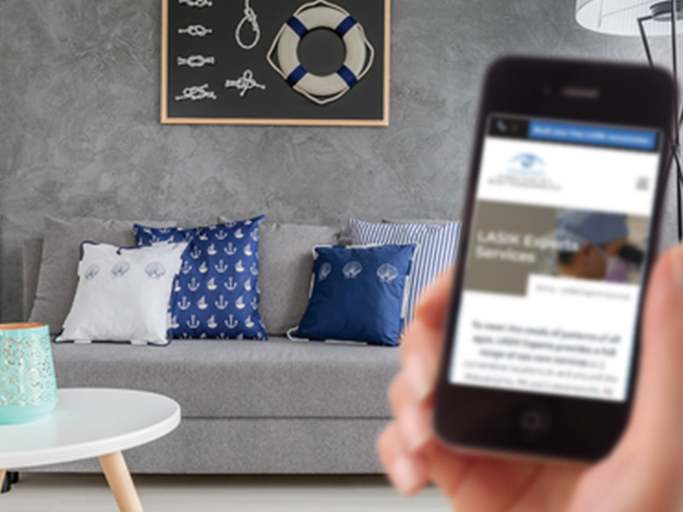 A right hand holding a phone with LASIK Experts website on the screen and a living room backdrop with couch and pillows. The phone is out of focus while the backdrop is in focus.