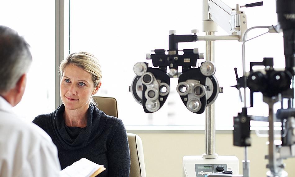 Inside an ophthalmologist’s office with eye examination equipment to the right. A female patient is seated towards the camera while a male doctor faces her